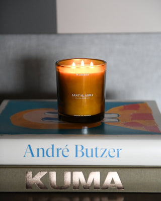 Vela Santal Aura 370 g de Madison en vidrio ámbar con tres pabilos encendidos, sobre libros de arte y arquitectura.